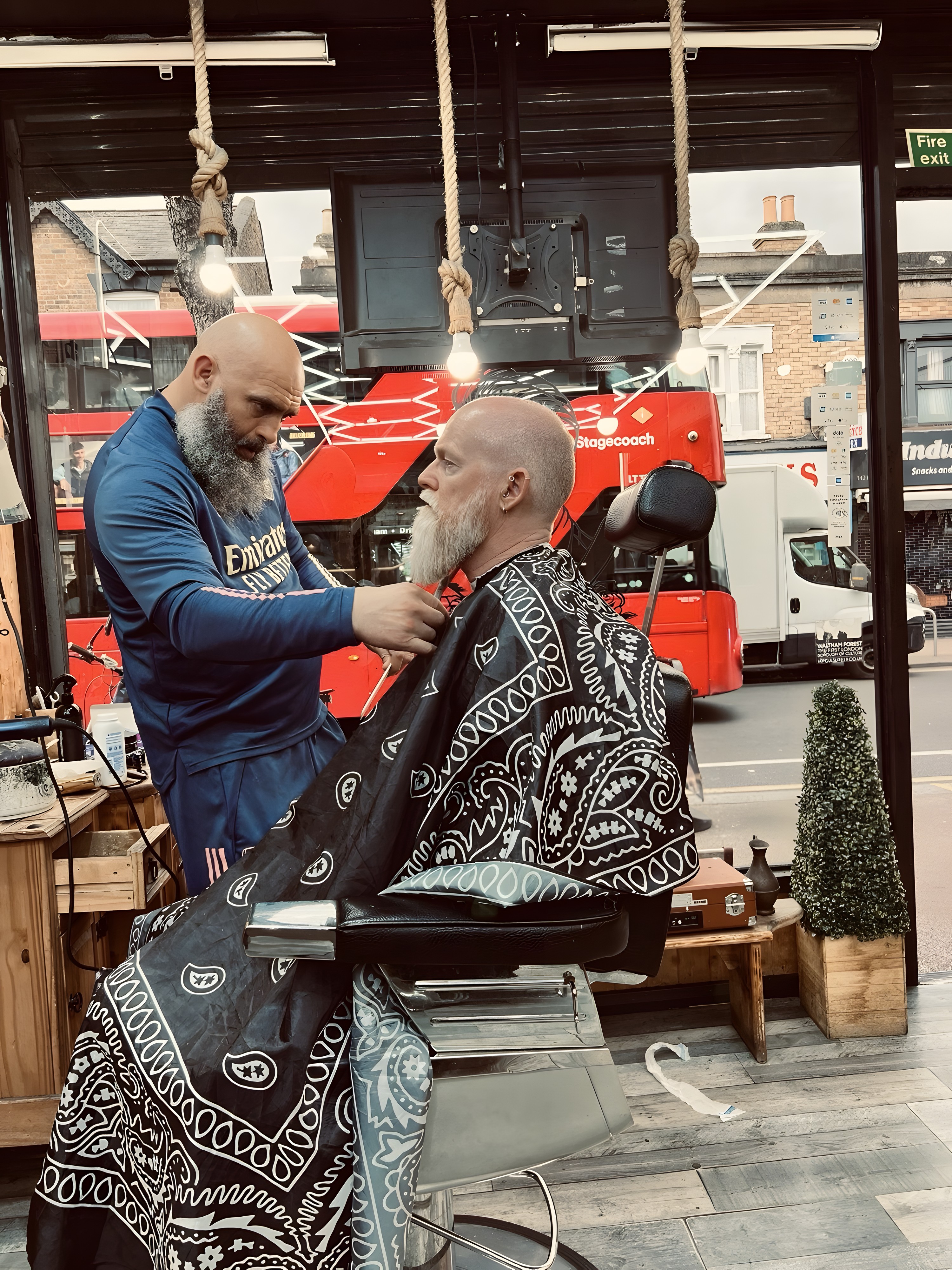 A barber at Vintage Cut trimming a customer's beard in the chair by the shopfront window, Hoe Street outside
